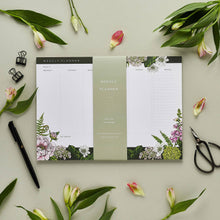 Load image into Gallery viewer, A styled photo of a weekly planner in the form of a pad that is meant to sit on a desk. The planner is illustrated with deep breen and purple florals along the bottom, and a hint of botanicals along the top. The planner is surrounded by flowers, a pen, some stationery clips, and a small pair of scissors
