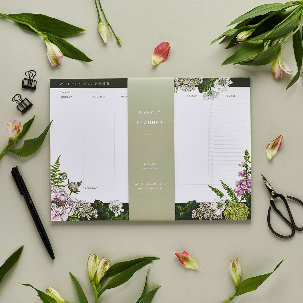 A styled photo of a weekly planner in the form of a pad that is meant to sit on a desk. The planner is illustrated with deep breen and purple florals along the bottom, and a hint of botanicals along the top. The planner is surrounded by flowers, a pen, some stationery clips, and a small pair of scissors