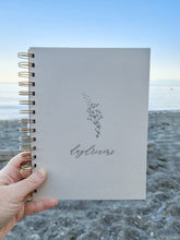 Load image into Gallery viewer, A picture of a beige or cream colored journal bound with copper coils. the word &quot;daydreams&quot; is written in calligraphy on the cover underneath a delicate botanical illustration. I hand is holding up the journal in nature, on a beach with a sunset, ocean, and sand backdrop.
