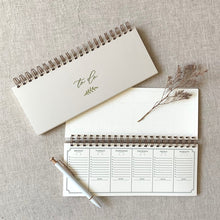 Load image into Gallery viewer, Two narrow rectangular shaped desk calendars, the legth of a computer keyboard, lay on a surface with a white pen and a twig, arranged so that one calendar displays the cover and another open to display the weekly calendar pages.  The front cover features forest green calligraphy and aand-drawn ferm, stamped with letterpress into high-quality matte board. The inner pages consist of rows for each day of the week with lines and checkboxes for 9 taks, plus a section below for taking notes. Copper bound h. 

