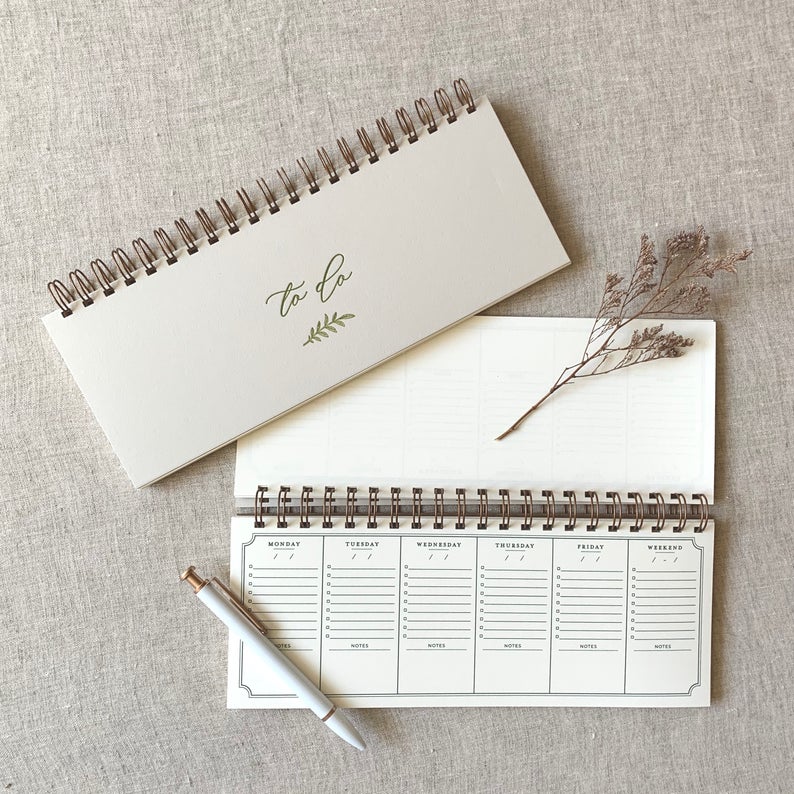 Two narrow rectangular shaped desk calendars, the legth of a computer keyboard, lay on a surface with a white pen and a twig, arranged so that one calendar displays the cover and another open to display the weekly calendar pages.  The front cover features forest green calligraphy and aand-drawn ferm, stamped with letterpress into high-quality matte board. The inner pages consist of rows for each day of the week with lines and checkboxes for 9 taks, plus a section below for taking notes. Copper bound h. 